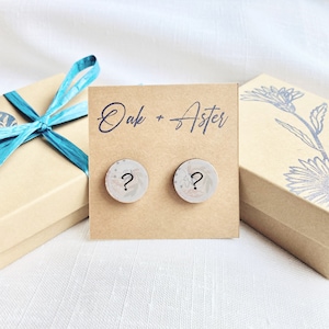 May include: A pair of wooden earrings with a floral design and a question mark on each earring. The earrings are displayed on a brown card with the text "Oak + Aster" written on it.