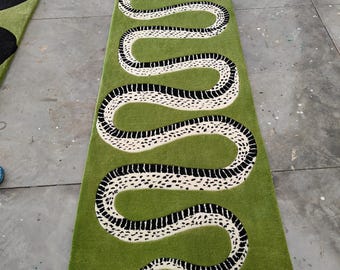 Puede incluir: Una alfombra pasillera larga y rectangular de color verde con un gráfico de una serpiente grande y sinuosa. La serpiente tiene un cuerpo blanco con manchas y rayas negras, y una pequeña lengua roja en la parte superior del diseño alargado.