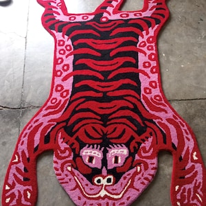 May include: A vibrant, handmade rug in the shape of a tiger. The rug features a red body with black stripes and pink accents. The tiger's face is detailed with pink, white, and black elements. The rug is designed to lay flat on the floor.