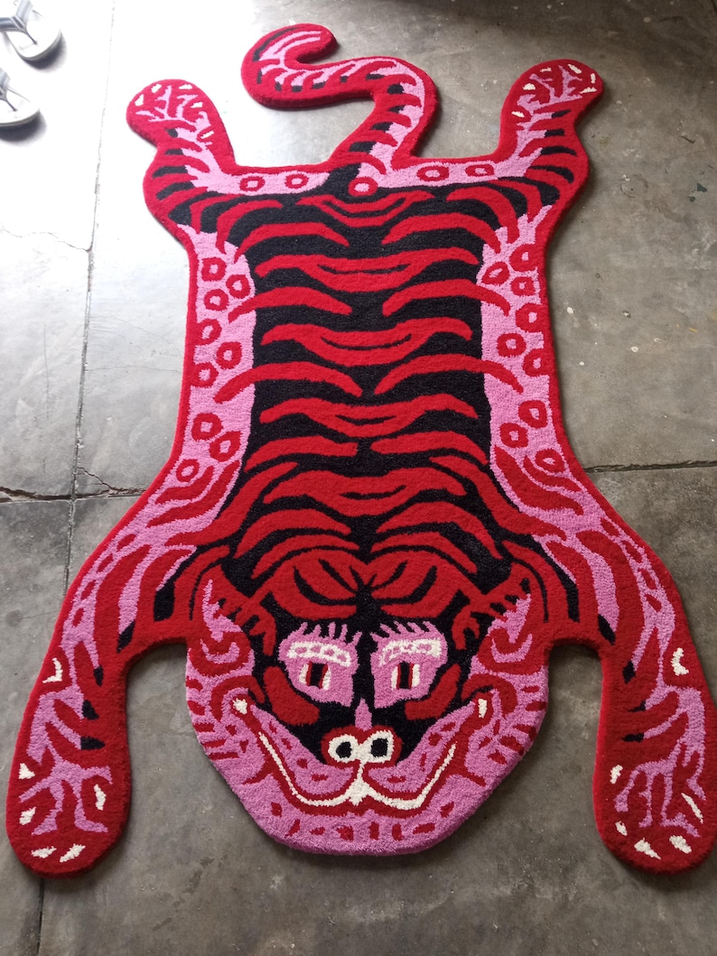 May include: A vibrant, handmade rug in the shape of a tiger. The rug features a red body with black stripes and pink accents. The tiger's face is detailed with pink, white, and black elements. The rug is designed to lay flat on the floor.