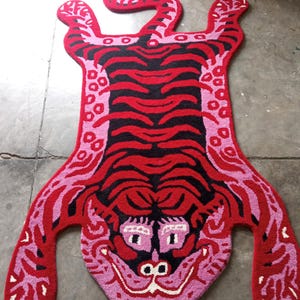 May include: A vibrant, hand-tufted rug in the shape of a tiger. The rug features a bold color palette of red, black, and pink, with a detailed face and striped body. The rug's dimensions are approximately 60 inches long.
