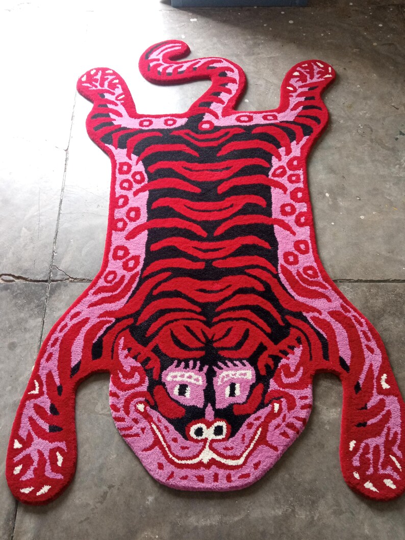 May include: A vibrant, hand-tufted rug in the shape of a tiger. The rug features a bold color palette of red, black, and pink, with a detailed face and striped body. The rug's dimensions are approximately 60 inches long.