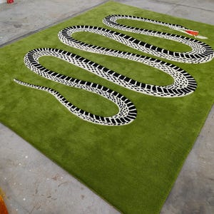 Può includere: Un tappeto verde con un grande disegno di serpente in bianco e nero, con una lingua rossa. Il tappeto ha una consistenza morbida e soffice, probabilmente in lana. Il disegno del serpente è centrale e il tappeto è rettangolare.
