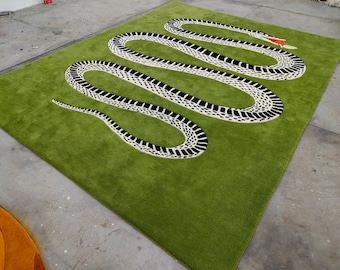Puede incluir: Una alfombra verde con un diseño de serpiente grande en blanco y negro, con una lengua roja. La alfombra tiene una textura suave y afelpada, probablemente de lana. El diseño de la serpiente es central y la alfombra es rectangular.