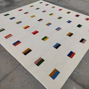 May include: A large, rectangular white rug with a repeating pattern of colorful rectangular blocks. The blocks are in various colors, including red, blue, yellow, and green. The rug is made of a soft, textured material.