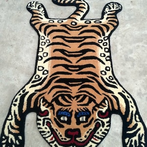 May include: A tiger-shaped rug with a tan and black striped design. The rug has a smiling face with blue eyes and red details. The rug is made from a soft, plush material, suitable for a bedroom or living room.