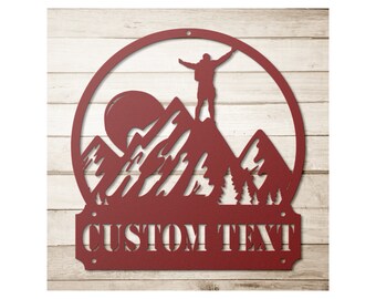 May include: A red metal wall art piece featuring a mountain scene with a person standing atop a peak with arms raised. The design includes a sun, trees, and the words "CUSTOM TEXT" at the bottom, all within a circular frame.