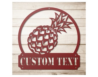 May include: Red metal pineapple wall art with a circular frame and a rectangular banner at the bottom. The banner reads "CUSTOM TEXT". The pineapple design is cut out, revealing the background. The art is mounted on a light wood-grain surface.