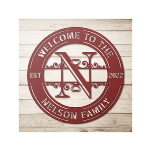 May include: A red metal family sign with the text "WELCOME TO THE NELSON FAMILY" and the year "2022". The letter "N" is prominently displayed in the center, with decorative flourishes and hearts. The sign is circular.