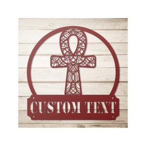 May include: A red metal wall sign featuring an Ankh symbol within a circle, with the words "CUSTOM TEXT" below. The Ankh has an intricate, openwork design. The sign is mounted on a light-colored, wooden plank background.