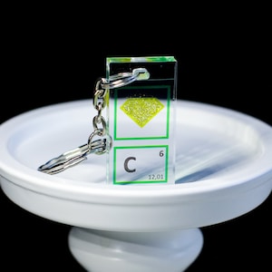 Può includere: Un portachiavi in acrilico trasparente con un bordo verde e un diamante giallo con glitter all'interno. Il portachiavi presenta anche il simbolo chimico del carbonio, "C", con il numero atomico "6" e il peso atomico "12,01".