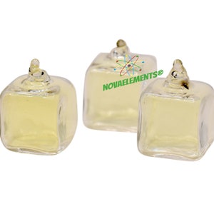 Puede incluir: Tres botellas de vidrio transparente con forma cuadrada y un pequeño tapón de vidrio transparente en la parte superior. Las botellas están llenas de un líquido amarillo claro. Las botellas están sobre una superficie blanca. El texto "NOVAELEMENTS®" es visible en la superficie.