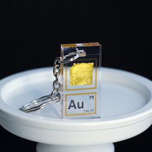 Può includere: Un portachiavi in oro con un ciondolo in acrilico trasparente che presenta l'elemento oro (Au) con il suo numero atomico (79) e il suo peso atomico (196,97).