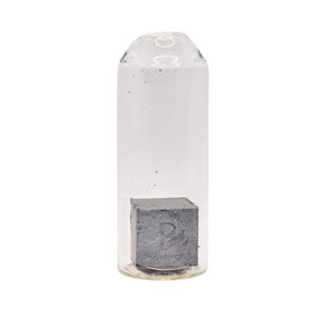 Puede incluir: Un vial de vidrio transparente que contiene un objeto cuadrado gris. El vial es cilíndrico con una parte superior redondeada. El objeto en el interior parece ser un cubo. El fondo es blanco liso.