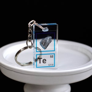 Può includere: Un portachiavi rettangolare trasparente con un portachiavi argentato. Il portachiavi presenta una grafica blu e bianca dell'elemento Tellurio (Te), numero atomico 52 e massa atomica 127,6, con un piccolo cristallo blu scuro all'interno.