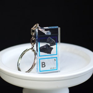 Op de afbeelding: Een transparante acryl sleutelhanger met een klein stukje boor element erin. De sleutelhanger heeft een zilveren metalen ring en de tekst "B 5 10.811" is onderaan gedrukt.