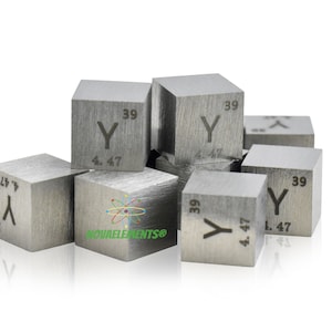 Puede incluir: Un grupo de pequeños cubos de color plateado, cada uno con el símbolo químico "Y" y el número atómico "39" grabado en un lado. Los cubos están dispuestos en una pila suelta sobre un fondo blanco.