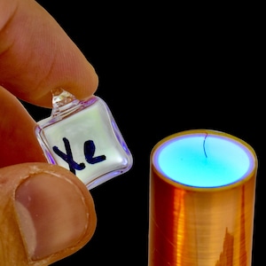 May include: A clear glass cube with the letter "Xe" written on it in black marker, held in a hand over a copper coil with a blue glow emanating from the top.