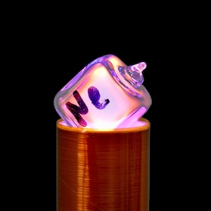 Puede incluir: Un tubo de vidrio transparente con la letra "Ne" en su interior, que brilla en púrpura, se encuentra sobre una bobina de cobre.