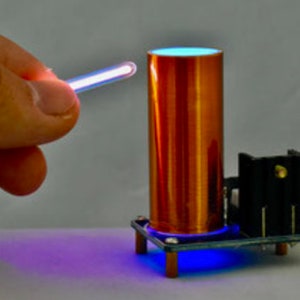 Puede incluir: Una bobina de cobre con un brillo azul en la parte superior, un tubo transparente que brilla en púrpura y una base negra con un brillo azul.