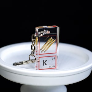 May include: A clear acrylic keychain with a silver chain. The keychain features a small square with the chemical symbol "K" for potassium, the atomic number "19", and the atomic weight "39.098". Inside the square, there are three thin gold wires.