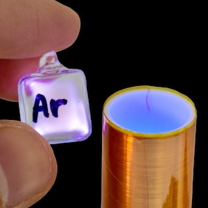 Puede incluir: Un vial de vidrio transparente lleno de un gas azul brillante, etiquetado como "Ar" para Argón, se sostiene en una mano sobre una bobina de cobre. La bobina emite un brillo azul.