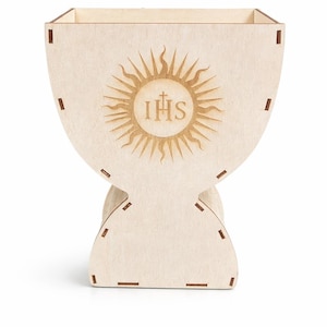 Puede incluir: Una caja de madera de color claro en forma de c&aacute;liz, con un dise&ntilde;o de sol y las letras IHS. La caja tiene una parte superior cuadrada y una base redondeada, con ranuras para el montaje. Decoraci&oacute;n de tem&aacute;tica religiosa.