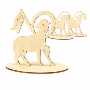 Peut inclure: Figurines en bois représentant des agneaux avec une auréole et un drapeau, posées sur une base ronde. L'agneau tient un drapeau avec une croix. D'autres figurines d'agneaux sont visibles en arrière-plan. Les figurines sont en bois clair.