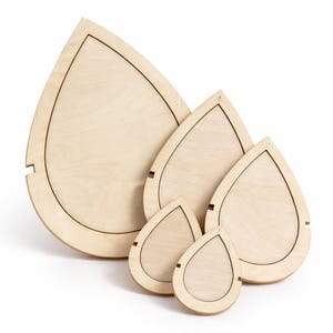 May include: A set of five wooden teardrop-shaped trays of varying sizes. Each tray has a light wood grain finish with a dark brown outline and a small notch on the side. The trays are arranged in a nested configuration.