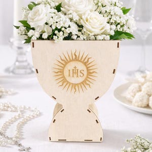 Puede incluir: Un jarr&oacute;n de madera en forma de c&aacute;liz con un dise&ntilde;o de sol y las letras IHS. Contiene rosas blancas y paniculata. Una vela, un rosario y un plato con dulces est&aacute;n en el fondo.