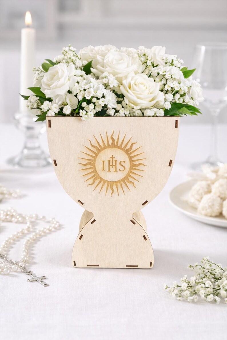 Puede incluir: Un jarr&oacute;n de madera en forma de c&aacute;liz con un dise&ntilde;o de sol y las letras IHS. Contiene rosas blancas y paniculata. Una vela, un rosario y un plato con dulces est&aacute;n en el fondo.