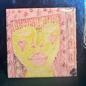 May include: A square tile featuring a stylized face with a yellow complexion, pink lips, and pink heart-shaped cheeks. The face is framed by a textured pink border with circular patterns and glittery accents throughout the entire surface.