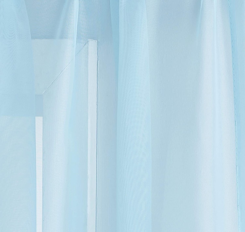 May include: A sheer light blue curtain panel hangs in a window. The curtain is made of a lightweight fabric that allows light to filter through.