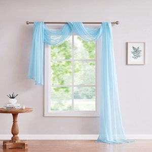 May include: A light blue sheer window scarf drapes over a window with white trim. The scarf is tied back on each side with a silver rod.