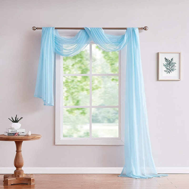 May include: A light blue sheer window scarf drapes over a window with white trim. The scarf is tied back on each side with a silver rod.