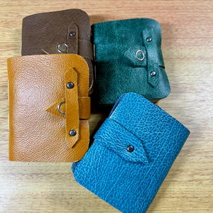 Op de afbeelding: Vier kleine leren portemonnees in tinten bruin, goud, groen en blauw. Elke portemonnee heeft een klepsluiting die is vastgezet met een metalen drukknop en een decoratieve metalen ring. De portemonnees zijn gerangschikt op een licht houten oppervlak.