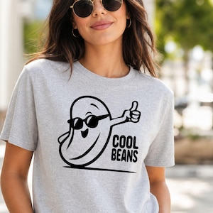 May include: A heather gray t-shirt featuring a black graphic of a smiling bean character wearing sunglasses and giving a thumbs up. The text COOL BEANS is printed in black capital letters next to the character on the front of the shirt.