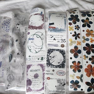 May include: Selection of decorative paper strips with vintage designs. Includes transparent sheets with stamps and text, and strips with floral and abstract patterns. The colour palette features muted tones of purple, brown, and cream.
