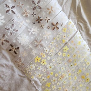 May include: A selection of washi tape rolls with floral patterns. The designs include flowers in brown, white, and yellow. The tape is semi-transparent and made of paper. The rolls are arranged side by side.