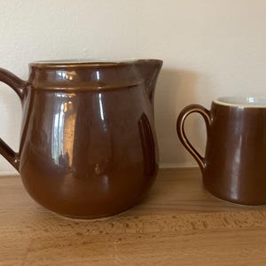 May include: Two brown ceramic jugs, one larger than the other. The larger jug has a handle and a rounded body. The smaller jug has a handle and a more cylindrical body.