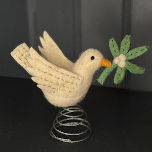 Op de afbeelding: Een crèmekleurig vilten duifbeeldje met een groen en witte bloem in zijn snavel. De duif heeft gouden stiksels en staat op een zilveren metalen veerbasis. De snavel van de vogel is oranje.