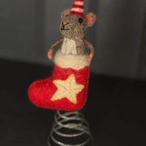 May include: A felt Christmas ornament featuring a grey mouse wearing a red and white striped hat, emerging from a red stocking with a gold star. The stocking sits atop a silver spring.