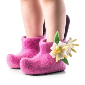 May include: Bright pink felt boots with a rounded toe and a decorative flower. The flower has white petals, yellow centres, and green leaves. The boots are designed for a child.