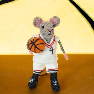 May include: A grey felt mouse wearing a white basketball jersey with red trim and the number 4. The mouse is holding an orange basketball.