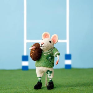 Könnte beinhalten: Eine handgefertigte Filzmaus-Figur in einem grünen Rugby-Outfit, die einen braunen Rugbyball hält. Die Maus trägt weiße Shorts mit einem grünen Kleeblatt, schwarze Schuhe und grün-weiß gestreifte Socken. Der Hintergrund zeigt einen blauen Himmel und Rugby-Pfosten.