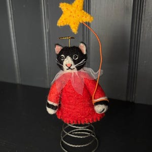 Puede incluir: Una caprichosa figura de gato con un atuendo de fieltro rojo, cuello blanco y una estrella amarilla sostenida por una cuerda naranja. El gato blanco y negro tiene un halo dorado y se asienta sobre una base de resorte plateado. Un artículo decorativo único.