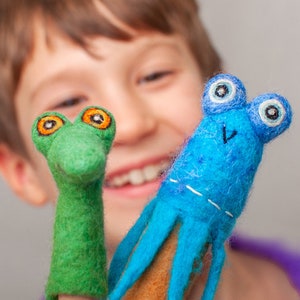 Könnte beinhalten: Zwei handgefertigte Fingerpuppen aus Filz. Eine ist grün mit orangefarbenen Augen und ähnelt einem Reptil. Die andere ist blau mit mehreren Tentakeln und großen Augen. Beide Puppen sind für fantasievolles Spielen und Geschichtenerzählen konzipiert.