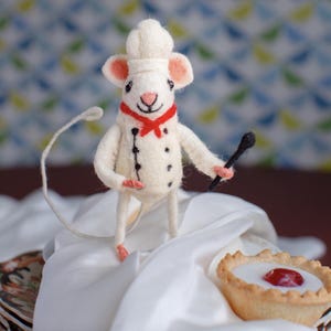 May include: A white felt mouse wearing a chef's hat and a red bow tie holds a black whisk. The mouse is standing on a white cloth next to a small pastry with a red cherry on top.