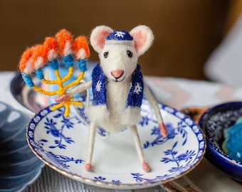 May include: A white felt mouse wearing a blue and white scarf and a blue hat holds a yellow menorah with orange and blue candles. The mouse is sitting on a white and blue patterned plate.