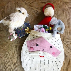 May include: A festive Christmas stocking shaped like Santa Claus, crafted from white felt with a pink nose and moustache. The stocking is filled with small gifts and toys, including a stuffed owl, a mouse, and a grey elephant. A red Santa hat is also present.